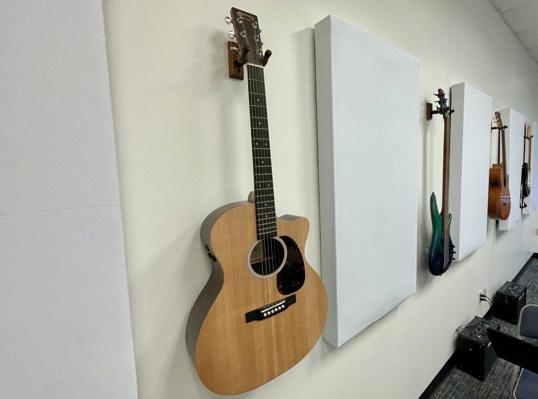 An acoustic guitar hanging on a practice room wall with electric guitars and amplifiers behind it
