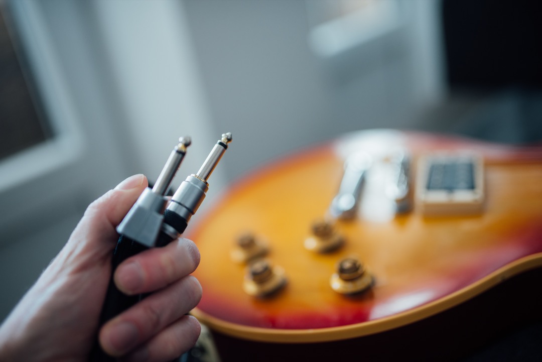 A hand holding two quarter-inch TS instrument jack plugs in front of a sunburst Les Paul style electric guitar