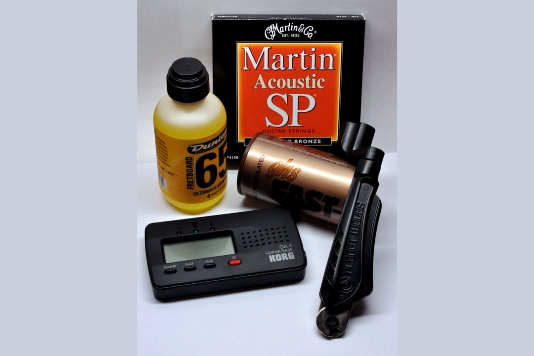 A guitar maintenance kit laid out on a pale background: a bottle of fretboard oil, a boxed set of acoustic strings, a string-cleaner tube, a clip-style chromatic tuner, and a string winder with built-in cutters